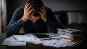 mężczyzna zestresowany długami siedzący przy stole z rachunkami i kalkulatorem w ciemnym pomieszczeniu